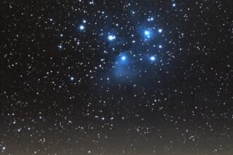 Star-filled night sky over a lit town on rolling hills, with a blue-trim cluster of stars near the top center.