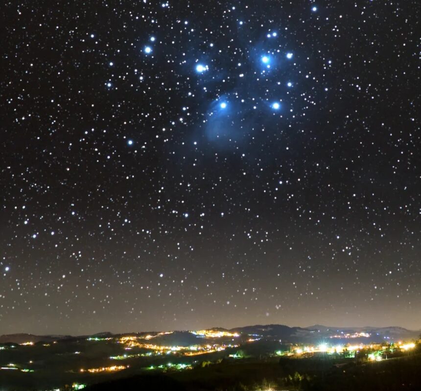 Star-filled night sky over a lit town on rolling hills, with a blue-trim cluster of stars near the top center.
