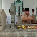 Man in brown uniform and woman in a hijab perform a blessing at a stone grave, with flowers laid on the tomb in a ceremonial setting.