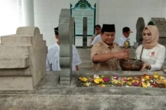 Man in brown uniform and woman in a hijab perform a blessing at a stone grave, with flowers laid on the tomb in a ceremonial setting.