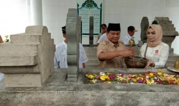 Man in brown uniform and woman in a hijab perform a blessing at a stone grave, with flowers laid on the tomb in a ceremonial setting.