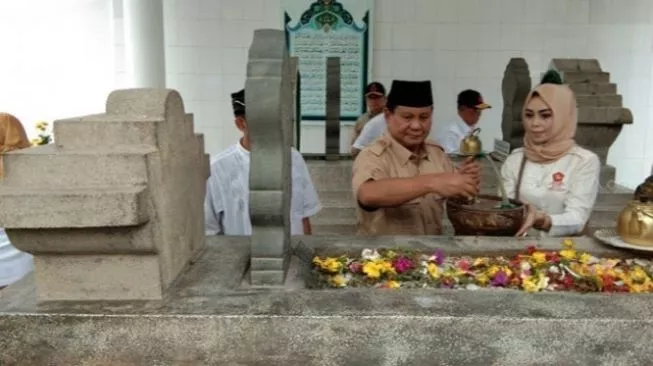 Man in brown uniform and woman in a hijab perform a blessing at a stone grave, with flowers laid on the tomb in a ceremonial setting.