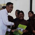Man in a white shirt shakes hands with a smiling recipient in a brown striped uniform while presenting a green certificate.