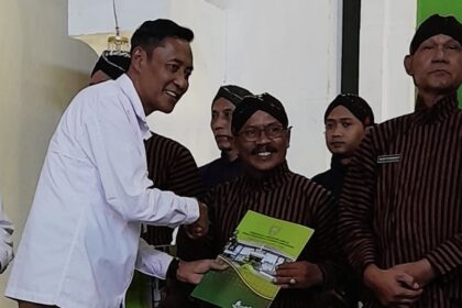 Man in a white shirt shakes hands with a smiling recipient in a brown striped uniform while presenting a green certificate.