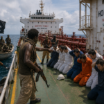 Armed pirates confront kneeling crew on a tanker deck; a black flag with skull and crossbones flutters nearby.
