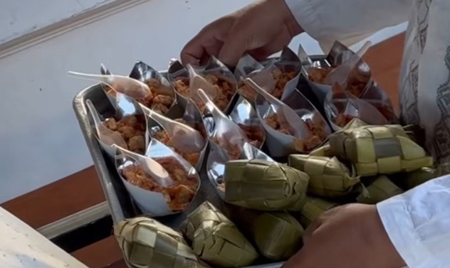 Tray of small foil cups filled with fried bite-sized pieces, each with a plastic spoon, arranged on a metal tray beside wrapped leaf packets held by a person in a white sleeve.