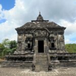 Front view of a dark stone temple with a central doorway, a broad staircase, and sculpted sides set in a park under a cloudy sky.