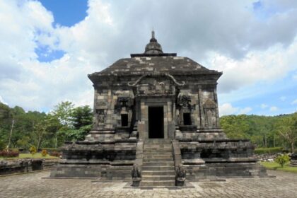 Front view of a dark stone temple with a central doorway, a broad staircase, and sculpted sides set in a park under a cloudy sky.