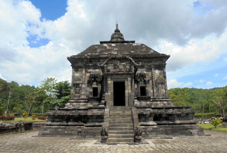 Front view of a dark stone temple with a central doorway, a broad staircase, and sculpted sides set in a park under a cloudy sky.
