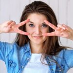 Young woman in a denim shirt making a playful peace sign around her eyes, against a light background.