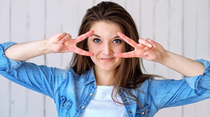 Young woman in a denim shirt making a playful peace sign around her eyes, against a light background.