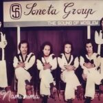 Nine-member band posing on a stage in matching white outfits with black trim; banner above reads 'Soneta Group' and subtitle 'The Sound of ...' as they sit and stand in a row.