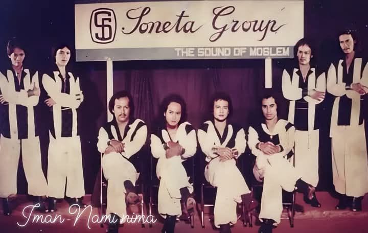 Nine-member band posing on a stage in matching white outfits with black trim; banner above reads 'Soneta Group' and subtitle 'The Sound of ...' as they sit and stand in a row.