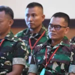 Four soldiers in camouflage uniforms sit indoors wearing red lanyards and ID badges, listening attentively.