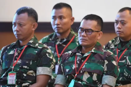 Four soldiers in camouflage uniforms sit indoors wearing red lanyards and ID badges, listening attentively.