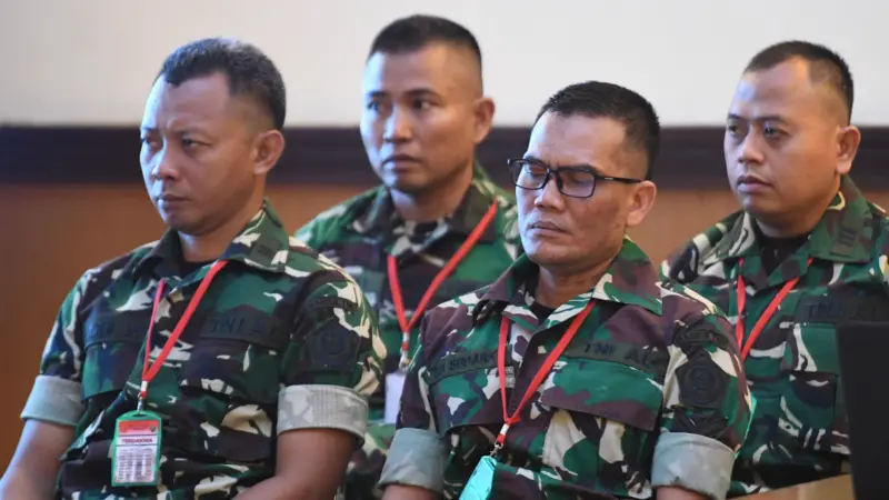 Four soldiers in camouflage uniforms sit indoors wearing red lanyards and ID badges, listening attentively.