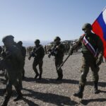 Soldiers in combat gear marching across a rocky desert, a large Russian flag carried by a soldier leads the group by a blue-sky backdrop.
