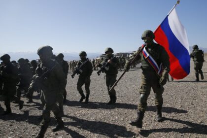 Soldiers in combat gear marching across a rocky desert, a large Russian flag carried by a soldier leads the group by a blue-sky backdrop.