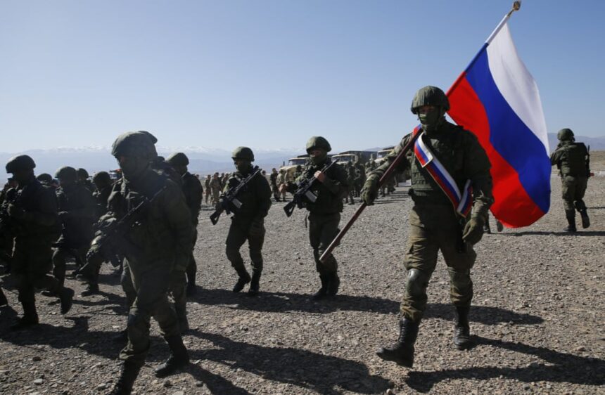 Soldiers in combat gear marching across a rocky desert, a large Russian flag carried by a soldier leads the group by a blue-sky backdrop.
