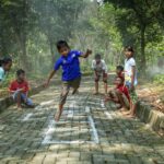 Children play hopscotch on a chalk-drawn square on a brick path in a forested area, with onlookers nearby.