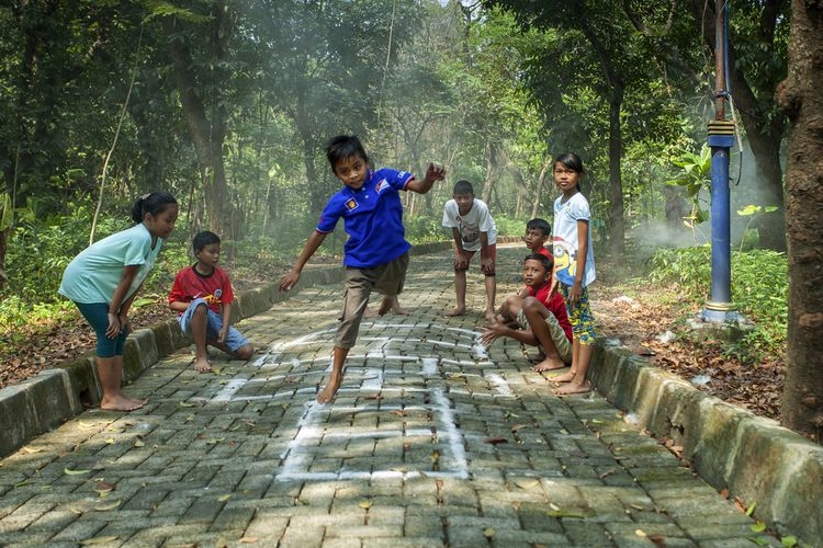 Children play hopscotch on a chalk-drawn square on a brick path in a forested area, with onlookers nearby.