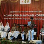 Students in batik and white hijabs sit on a stage during a cooperative creativity event; red backdrop reads 'AJANG KREASI INOVASI KOPERASI' with logos above.