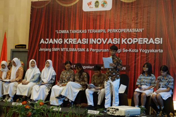 Students in batik and white hijabs sit on a stage during a cooperative creativity event; red backdrop reads 'AJANG KREASI INOVASI KOPERASI' with logos above.