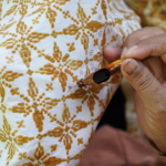 Close-up of a hand holding a small needle near brown embroidered floral pattern on white fabric, illustrating sewing detail