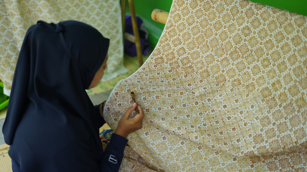Person in a dark blue hijab sewing a beige-brown patterned fabric by hand, close to the fabric's edge on a sewing frame.