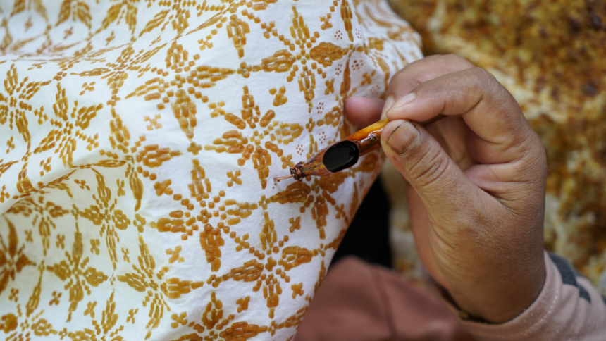 Close-up of a hand holding a small needle near brown embroidered floral pattern on white fabric, illustrating sewing detail
