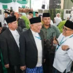 Group of men in peci hats and suits talking at a formal ceremony, with a crowd in green and white outfits behind them.