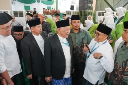 Group of men in peci hats and suits talking at a formal ceremony, with a crowd in green and white outfits behind them.