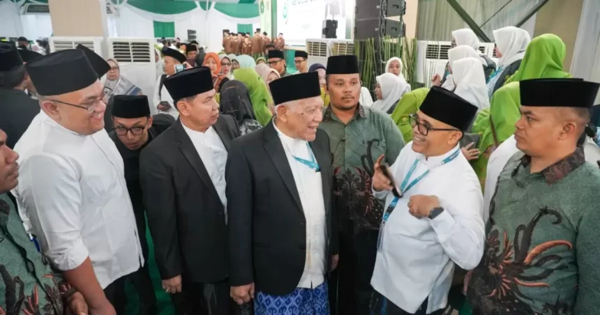 Group of men in peci hats and suits talking at a formal ceremony, with a crowd in green and white outfits behind them.