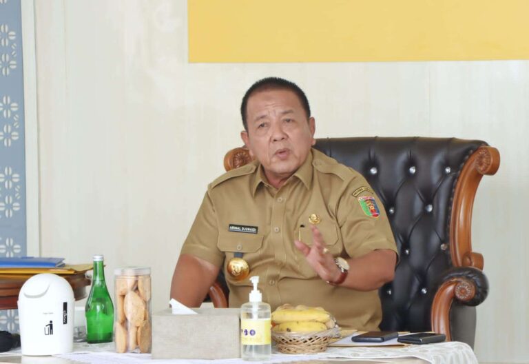 Man in a tan uniform seated at a desk, gesturing with his right hand as he speaks.