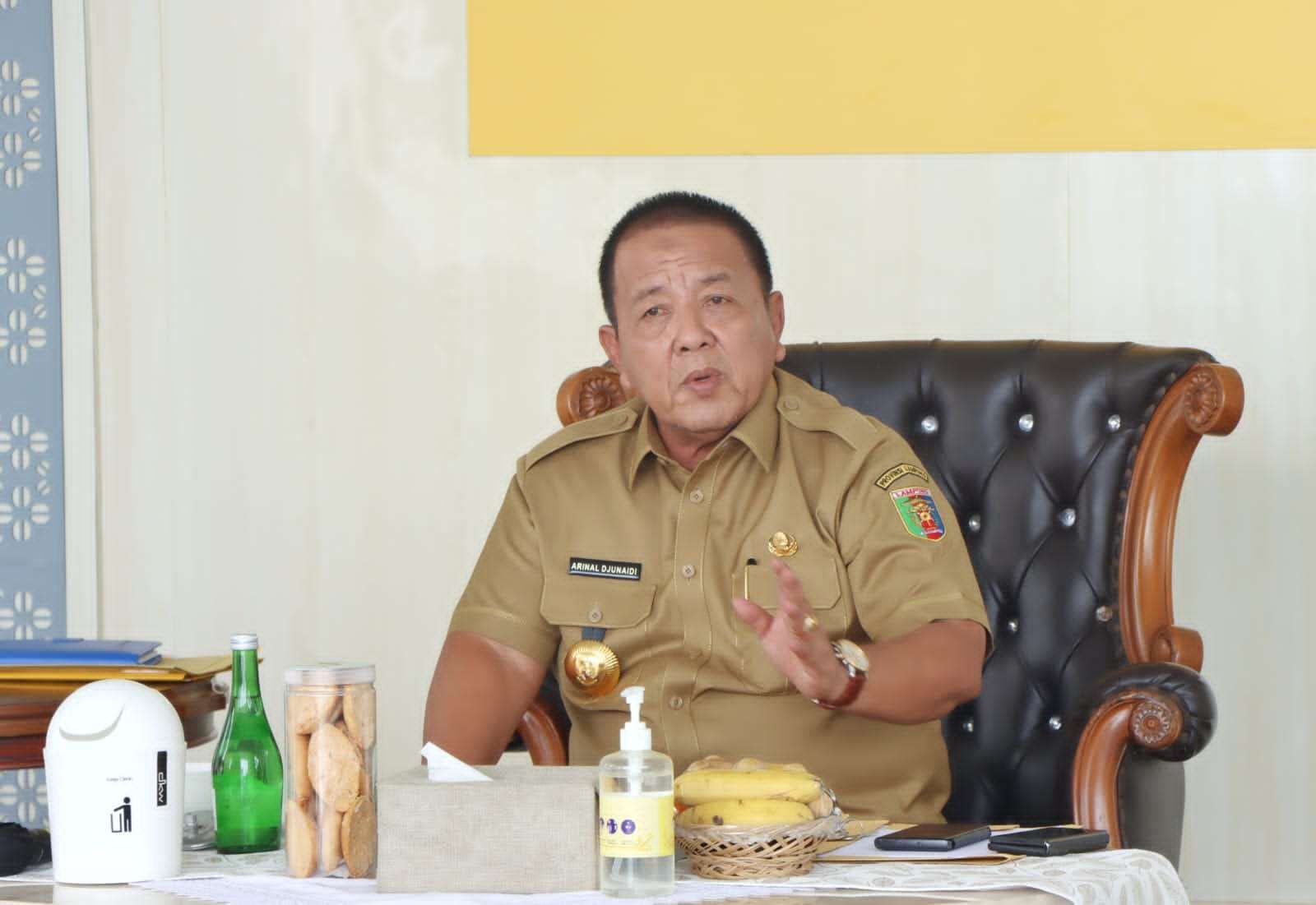 Man in a tan uniform seated at a desk, gesturing with his right hand as he speaks.