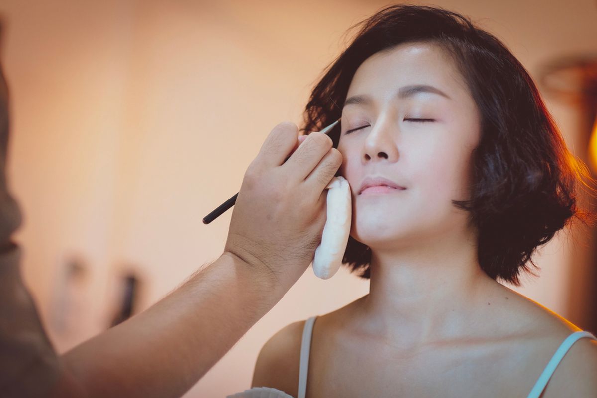 Makeup artist applies eye makeup to a woman with eyes closed and short dark hair.