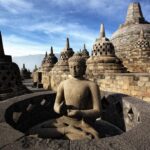 Buddha statue meditating inside a circular stone shrine at Borobudur, surrounded by bell-shaped stupas under a blue sky.
