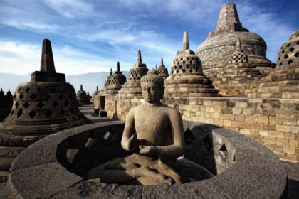 Buddha statue meditating inside a circular stone shrine at Borobudur, surrounded by bell-shaped stupas under a blue sky.