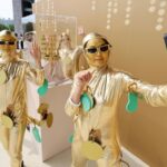 Three people in shiny gold outfits pose and selfie with a smartphone against a gold wall backdrop.