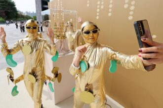 Three people in shiny gold outfits pose and selfie with a smartphone against a gold wall backdrop.