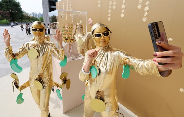 Three people in shiny gold outfits pose and selfie with a smartphone against a gold wall backdrop.