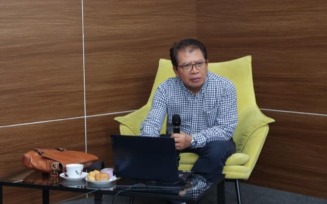 5twresfd - Mabur.co Man in a checkered shirt seated in a yellow armchair, speaking into a handheld microphone with a laptop on a glass table.