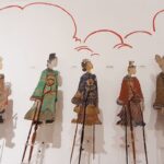 Five intricately dressed marionette puppets on tall stilts stand against a white wall with red cloud drawings in a museum display.