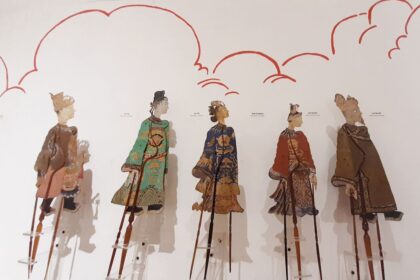Five intricately dressed marionette puppets on tall stilts stand against a white wall with red cloud drawings in a museum display.