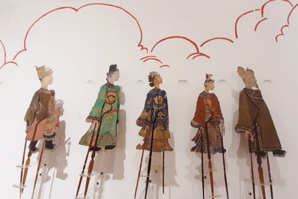 Five intricately dressed marionette puppets on tall stilts stand against a white wall with red cloud drawings in a museum display.