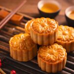 Mooncakes with flower-patterned tops stacked on a wooden surface with tea cups in the background.