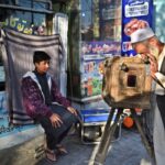 Elderly man in traditional dress peers through an old wooden box camera on a tripod as a boy sits nearby near a storefront with colorful signs.