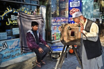 Elderly man in traditional dress peers through an old wooden box camera on a tripod as a boy sits nearby near a storefront with colorful signs.