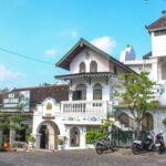 White colonial-style building with arches and balconies, motorcycles parked along the curb on a sunny street.