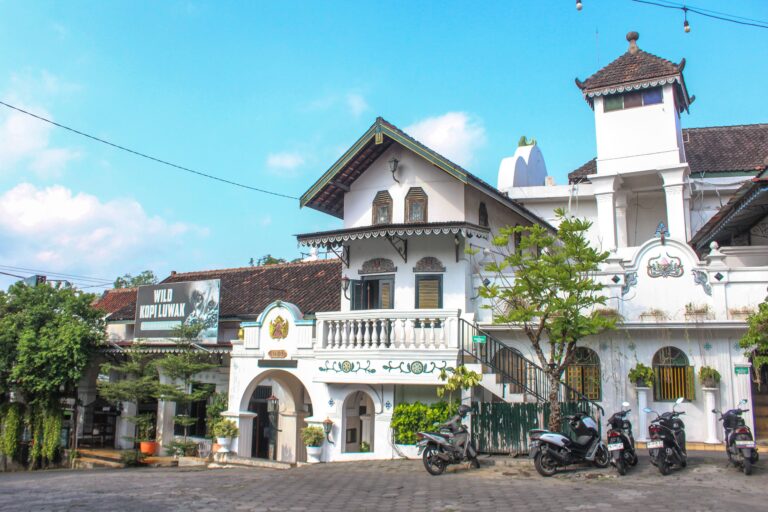 61dae8238722c9d48870e75295aae2f3 - Mabur.co White colonial-style building with arches and balconies, motorcycles parked along the curb on a sunny street.